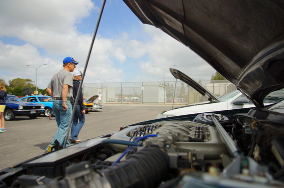 Engine bay with hood open, showing engine components. Other cars visible in background