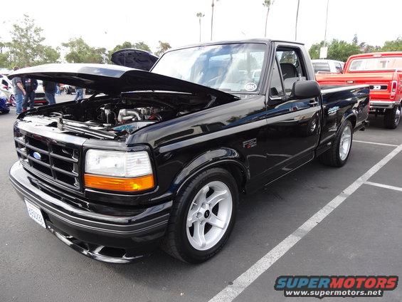 Black 1994 Ford F-150 pickup truck, hood open, showing engine. Another red truck is parked behind it