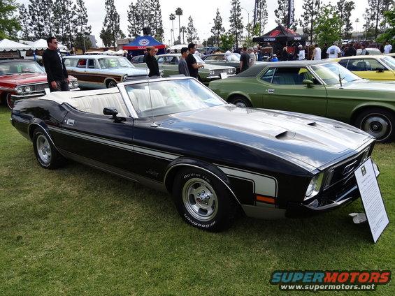 Black 1971 Ford Mustang convertible with white interior and silver racing stripes on hood and sides, parked on grass with other cars and tents in background