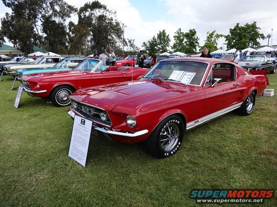 Red classic car with white stripe on side, parked on grass at car show. Other classic cars visible