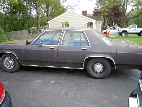 Dark brown Ford sedan parked in driveway, side view. Another truck and car are visible in the background