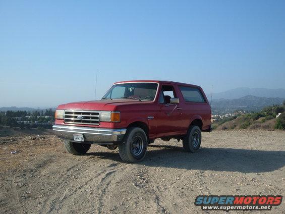 Red Ford Bronco parked on a dirt road with a town and hills in the background