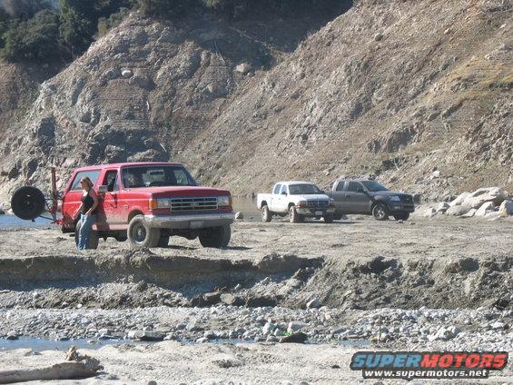 Red Ford Bronco parked by a rocky shore with a woman leaning on it; two other trucks are in the background