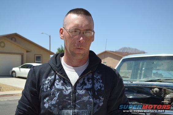 Man with buzz cut and glasses stands in front of a light blue truck with the hood open. A white car is parked in the driveway behind him