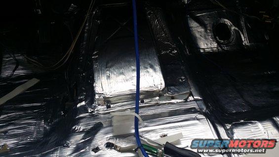 Interior of a vehicle with silver and black soundproofing material covering the floor and lower walls, with wires and a blue tube visible