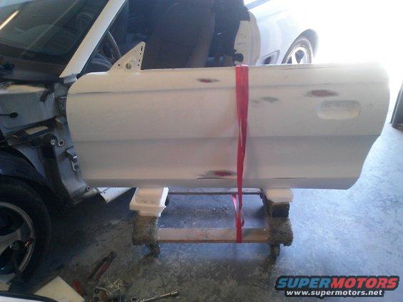Car door, possibly primed, held by red straps on a wooden stand