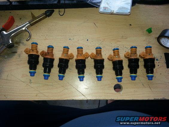 Eight fuel injectors laid out on a wooden surface next to an air duster