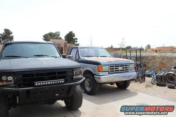 Two trucks parked outside, one black with a light bar, the other blue and silver with rust. Bicycles and a motorcycle are nearby