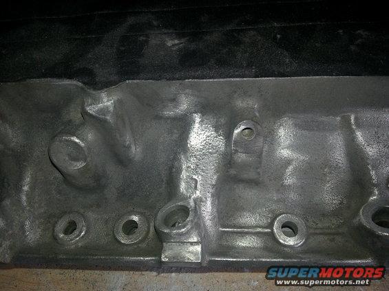 Close-up of a metal engine block with bolt holes and cast details. Supermotors.net logo visible
