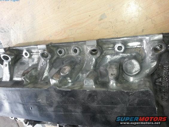 Close-up of a metal engine cylinder head with bolt holes and casting marks. Part number visible