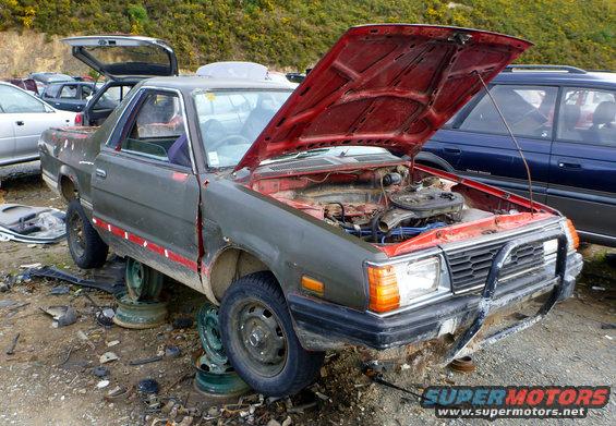 Gray Subaru Brat with hood open, missing wheels, in junkyard