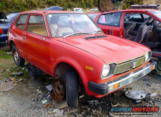 Red hatchback car with missing wheels in junkyard, another red car visible behind it