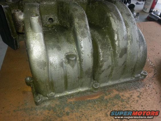 Aluminum intake manifold with green residue, resting on a brown surface with debris