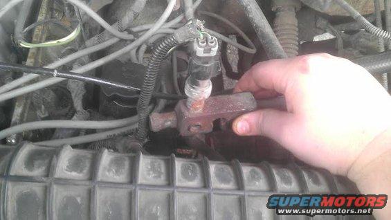Hand touching a sensor in a vehicle's engine bay, with wires and hoses visible
