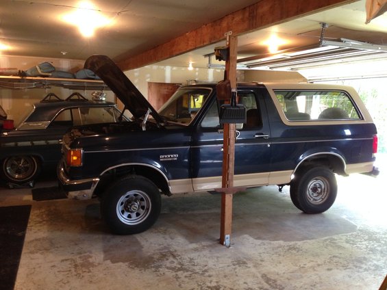 Dark blue and tan 1990 Ford Bronco with hood open, parked in a garage next to a classic car