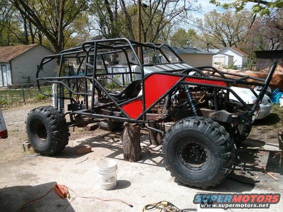 Custom buggy frame with large tires, red panels, and roll cage, sitting on blocks in a driveway