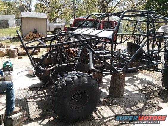 Off-road buggy frame on blocks, with large tires and exposed engine components. A red pickup truck is visible behind it
