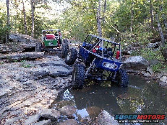 Two custom off-road buggies driving through a rocky stream bed. Blue buggy in foreground, green buggy behind it