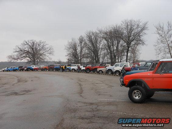 Many Ford Broncos parked in a line on a cloudy day. Orange Bronco in foreground