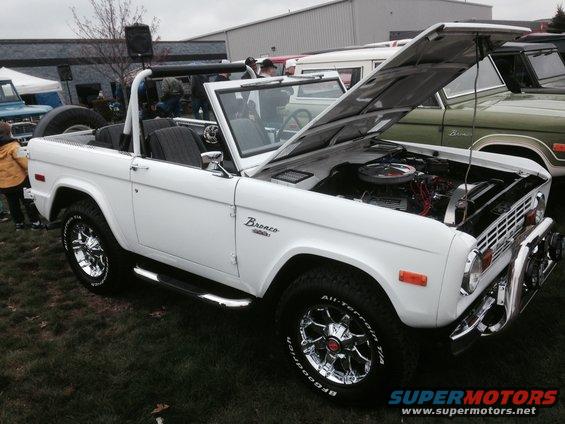 White 1970s Ford Bronco convertible, hood open, chrome wheels, side steps, parked on grass