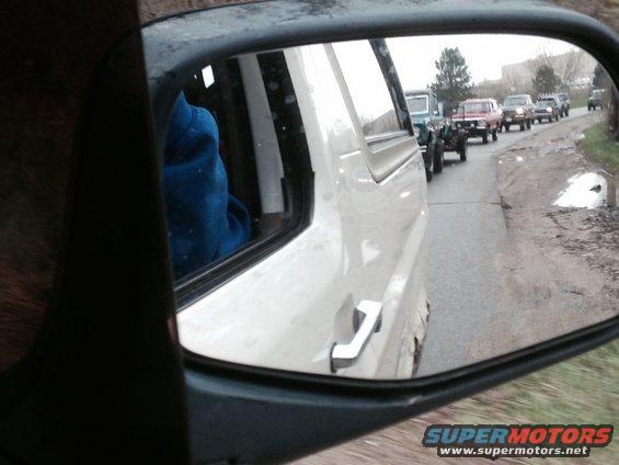 White pickup truck side mirror reflection shows a line of older SUVs and trucks on a wet road