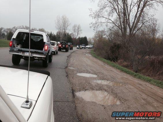 Rear view of a white 1995 Ford Bronco with open tailgate, leading a line of SUVs on a dirt road