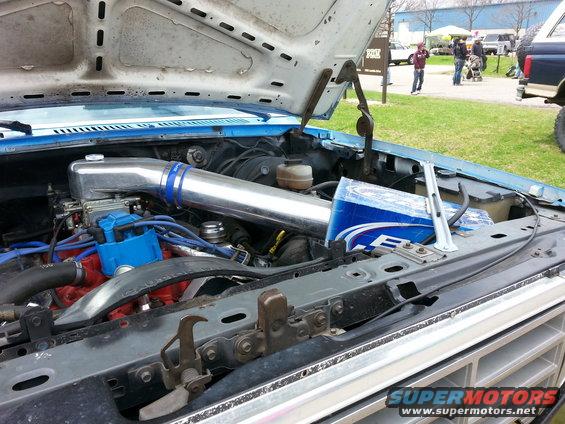 Engine bay with shiny intake tube and blue distributor cap. Some people and other vehicles in background