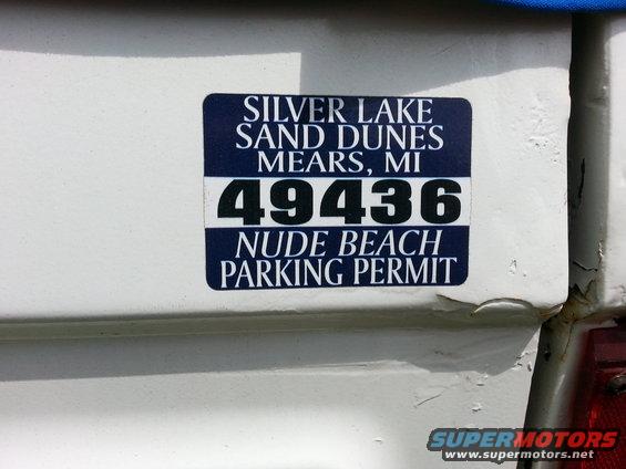 White vehicle exterior with a dark blue sticker reading 'SILVER LAKE SAND DUNES MEARS, MI 49436 NUDE BEACH PARKING PERMIT'