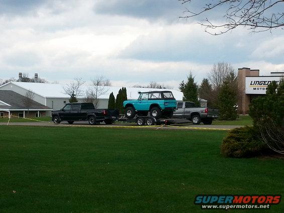 Blue Ford Bronco on a trailer towed by a black pickup truck, with a silver pickup truck behind it. Building in background