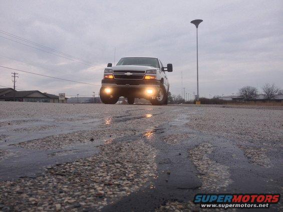White 2006 Chevrolet Silverado 2500 truck, front view, with headlights on, on a wet road