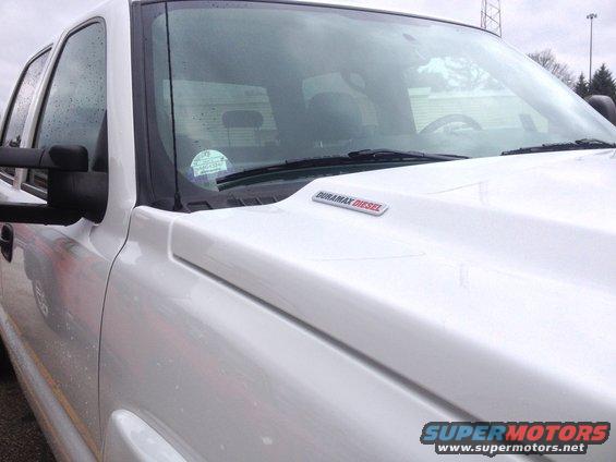 White pickup truck, hood has a Duramax Diesel badge. Windshield has a sticker