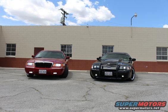Red sedan and black police-style sedan parked outside a building. Red sedan has 'EVIL MGM' plate. Black sedan has push bar and 'EVIL VTC' plate