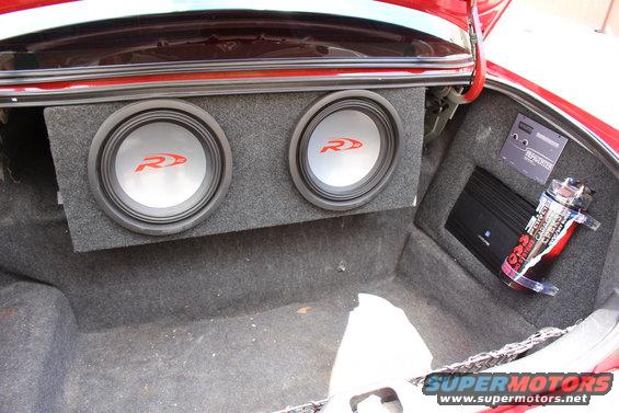 Car trunk interior with dual Alpine subwoofers in a gray carpeted box, an amplifier, and a capacitor