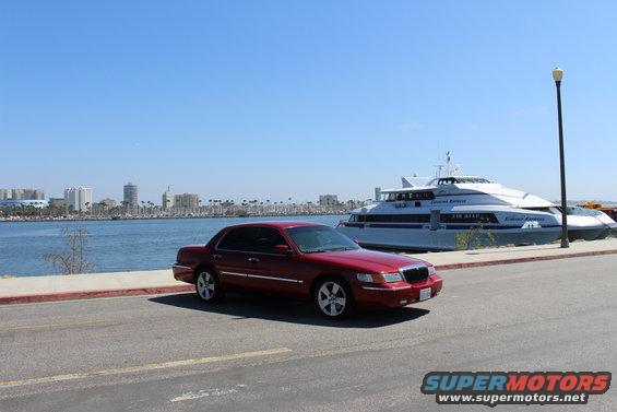 Red 2002 Mercury Grand Marquis parked by a waterfront with a large white boat and city skyline in the background