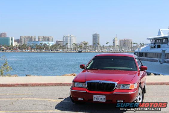 Red 2002 Mercury Grand Marquis parked by the water with a city skyline and a large boat in the background