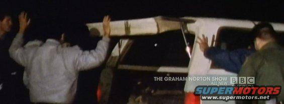 People with hands up near the rear of a dark SUV with a white roof rack