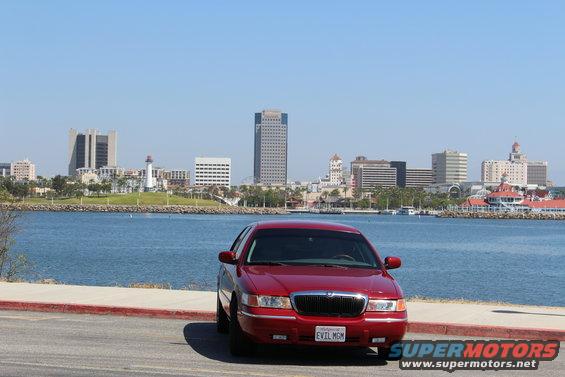 Red 2002 Mercury Grand Marquis parked facing front with a city skyline and water in the background. License plate reads EVIL MGM