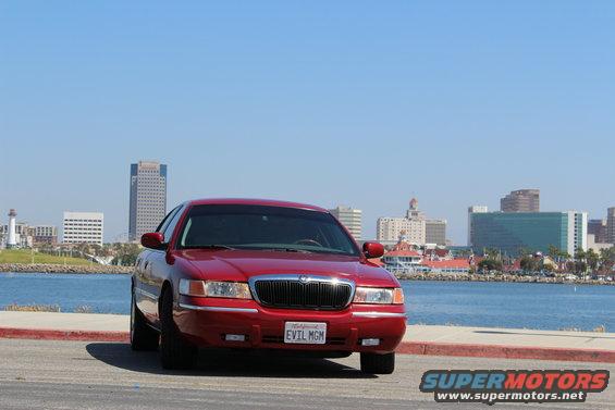 Red 2002 Mercury Grand Marquis with license plate "EVIL MGM" parked facing front, waterfront and city skyline behind