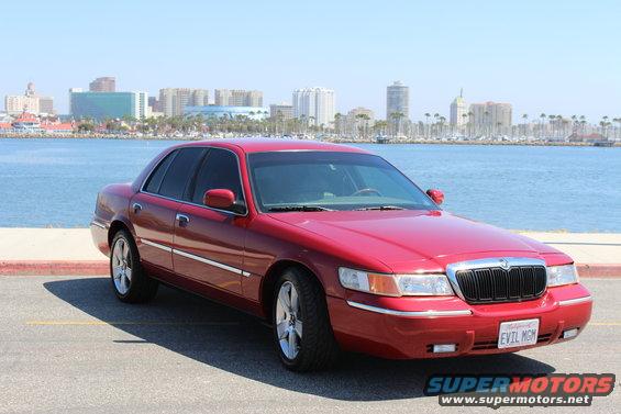 2002 Mercury Grand Marquis parked by the water, city skyline behind it. License plate reads 'EVIL MGM'