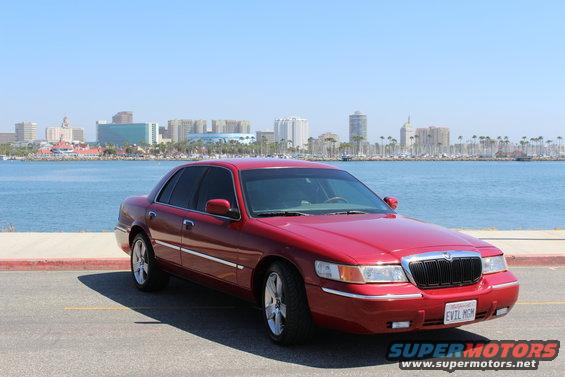 Red 2002 Mercury Grand Marquis parked by a waterfront with a city skyline in the background