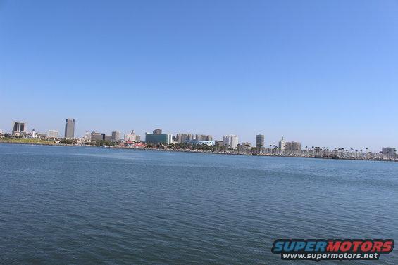 City skyline with buildings and palm trees visible across a wide expanse of blue water under a clear sky. Watermark reads SUPERMOTORS
