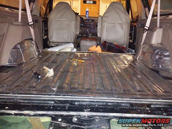 Interior of a vehicle with seats removed, showing soundproofing material on the floor and wheel wells. Tools are on the floor