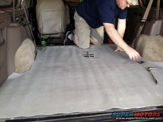 Man installing soundproofing material on the floor of an SUV's cargo area. Seats are visible in the background