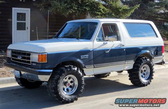 Two-tone blue and white Ford Bronco with large off-road tires parked in front of a house
