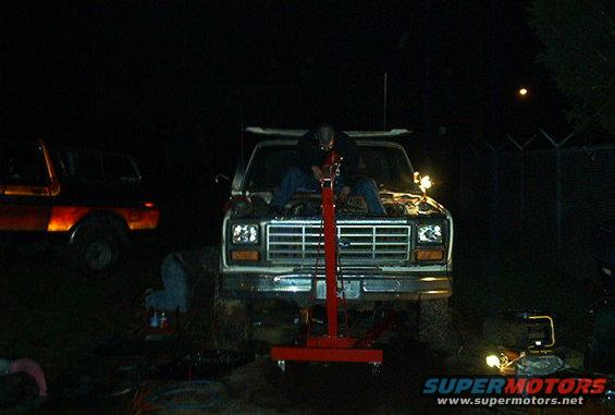 Ford truck with hood open, person working on engine with red lift. Another truck visible to the left
