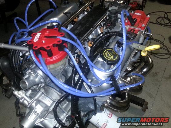 Ford V8 engine block with MSD distributor, spark plug wires, and exhaust headers. Oil fill cap visible
