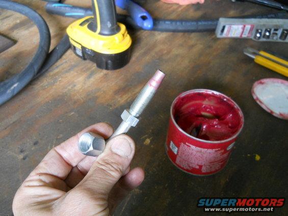 Hand holding a threaded rod with a nut, next to an open can of red grease. Tools and a work surface are in the background