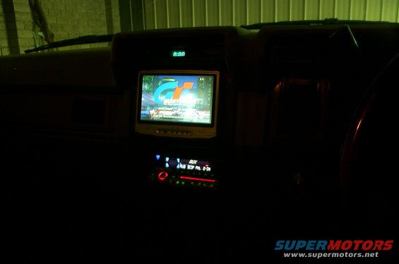 Car dashboard showing a screen displaying 'GT' logo and an aftermarket stereo below. Digital clock reads 0:00
