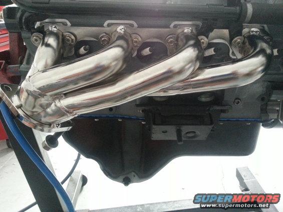 Chrome exhaust manifold on an engine block, with blue hose and Supermotors.net logo
