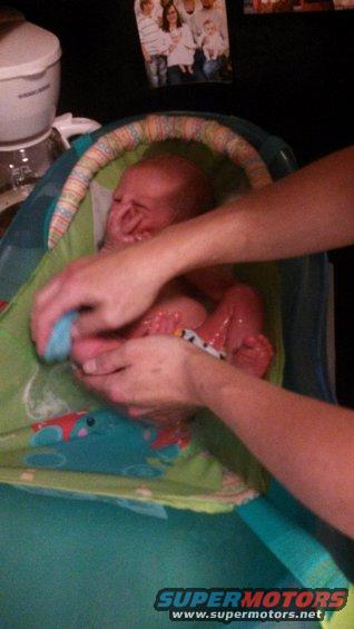 Baby in a bath seat with hands washing it. Family photo on the wall in background